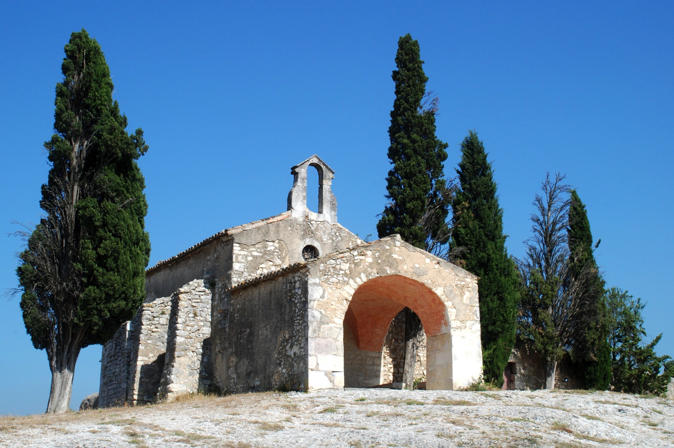 Old stone chapel with a large arched entrance, surrounded by tall cypress trees under a clear blue sky.